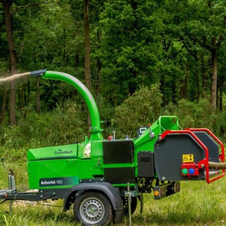 Arborist 150 6″ Road Towed Wood Chipper A green wood chipper machine, available through tool hire Newry, processes branches in a forested area, ejecting wood chips through an overhead chute.