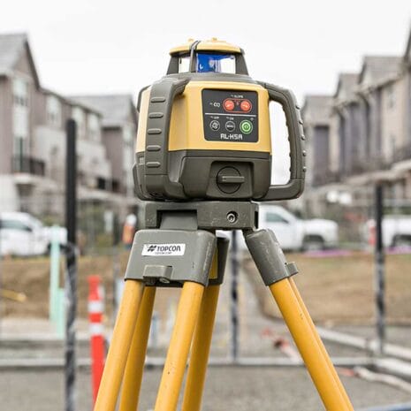 Topcon RL-H5A Rotating Laser Level A yellow and grey laser level on a tripod, available from tool hire Newry, is set up at a construction site with buildings in the background.