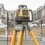 A yellow and grey laser level on a tripod, available from tool hire Newry, is set up at a construction site with buildings in the background.