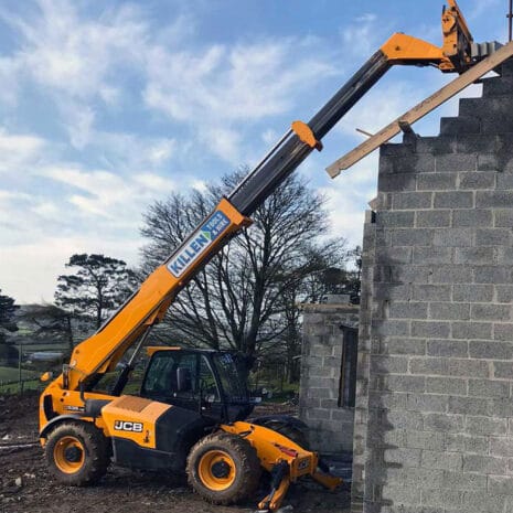 A yellow JCB telehandler from tool hire Newry, with an extended boom, lifts a wooden beam to the upper level of a partially constructed grey brick building.