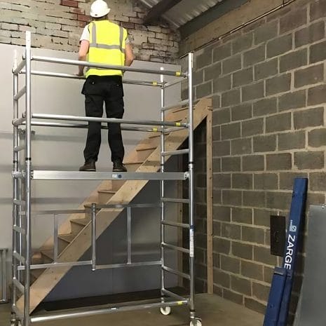 Aluminium Scaffold A construction worker in a hi-vis vest stands on an aluminium scaffold, hired from tool hire Newry, facing a wall near a staircase in an unfinished building.