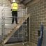 A construction worker in a hi-vis vest stands on an aluminium scaffold, hired from tool hire Newry, facing a wall near a staircase in an unfinished building.