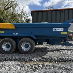 Kane Halfpipe Dump Trailer-2 A blue Kane Halfpipe Dump Trailer with yellow detailing, available from Tool Hire Newry, is parked on gravel beside a metal industrial building under a partly cloudy sky.