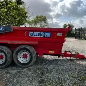 A Tuff Mac DT14 Dump Trailer from Killen Tools & Hire, your trusted tool hire in Newry, is parked on gravel near trees and a tractor under a cloudy sky.