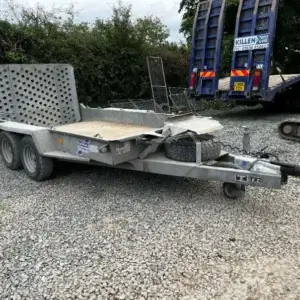 A damaged Ifor Williams plant trailer with a bent front and flatbed is parked on gravel near construction equipment and a hedge at a tool hire Newry location.