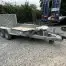 A damaged Ifor Williams plant trailer with a bent front and flatbed is parked on gravel near construction equipment and a hedge at a tool hire Newry location.