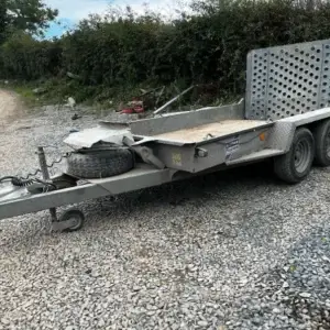 IFor Williams Plant Trailer-2 An Ifor Williams Plant Trailer with twin axles, a spare tyre, and a perforated loading ramp is parked on gravel near a dirt road and bushes—ideal for tool hire Newry customers needing reliable transport solutions.