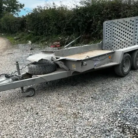 IFor Williams Plant Trailer-2 An Ifor Williams Plant Trailer with twin axles, a spare tyre, and a perforated loading ramp is parked on gravel near a dirt road and bushes—ideal for tool hire Newry customers needing reliable transport solutions.