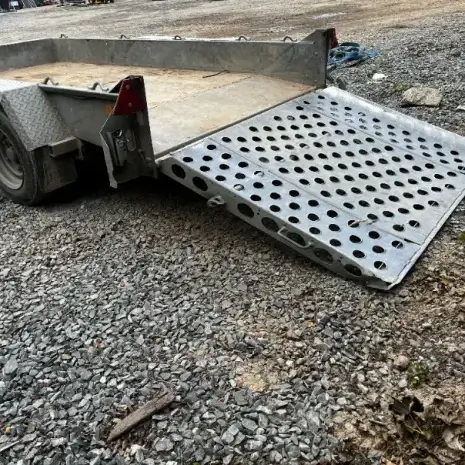 IFor Williams Plant Trailer-4 The Ifor Williams Plant Trailer, a metal twin-axle model with a perforated ramp lowered onto gravel, is ready for loading—perfect for transporting equipment from Tool Hire Newry.