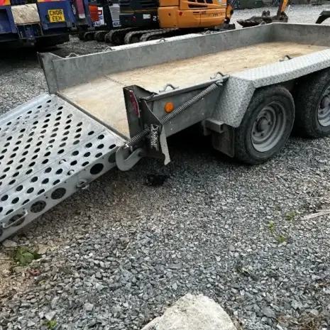 IFor Williams Plant Trailer-5 An Ifor Williams Plant Trailer, featuring twin axles and a perforated loading ramp, is parked on gravel at a work site. Available through tool hire Newry.