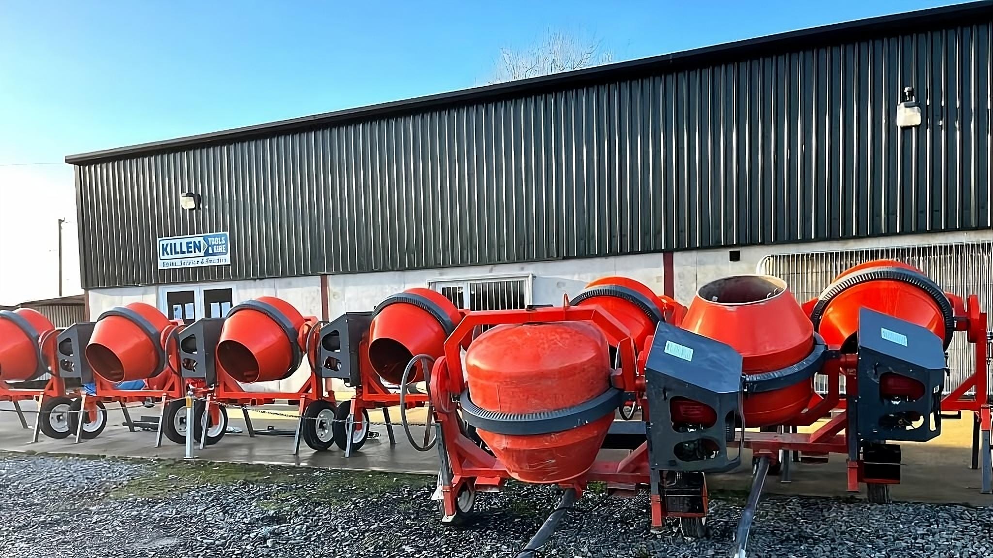 A row of red cement mixers is lined up outside a building with a corrugated metal exterior and a "KILEN" sign, suggesting this could be a tool hire Newry location.