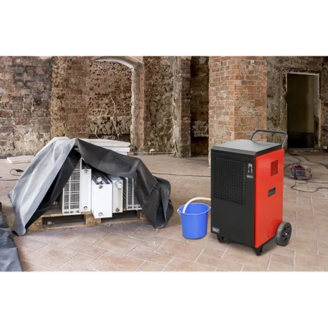 Sealey 70L Industrial Dehumidifier SDH70-4 The Sealey 70L Industrial Dehumidifier SDH70, available at tool hire Newry, is placed on a tiled floor beside a blue bucket and covered equipment in a partially renovated room with exposed brick walls.