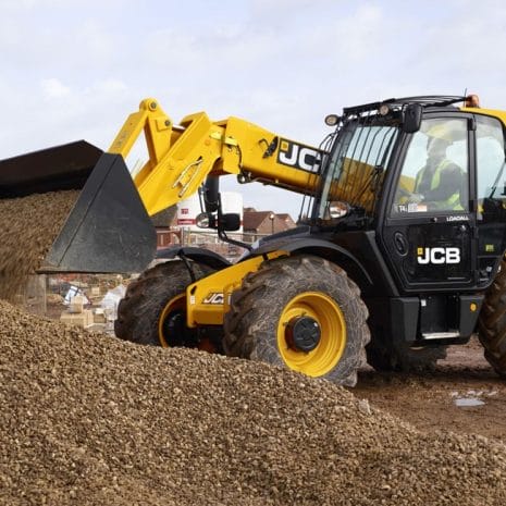 531-70 Telescopic handler-3 A yellow 531-70 telehandler from tool hire Newry moves a pile of gravel at a building site, with buildings visible in the background.