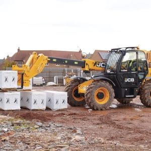 A yellow 531-70 Telescopic handler from tool hire Newry lifts a pallet of white construction blocks at a building site, with houses and materials visible in the background.