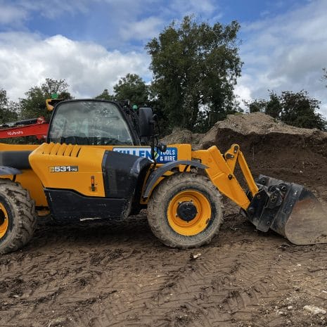 531-70 Telescopic handler-4 A yellow 531-70 telescopic handler with a front bucket attachment is parked on a dirt construction site, surrounded by soil heaps and treesโperfect for tool hire Newry projects.