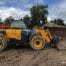 A yellow 531-70 telescopic handler with a front bucket attachment is parked on a dirt construction site, surrounded by soil heaps and treesโperfect for tool hire Newry projects.