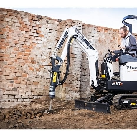 Bobcat E10Z Mini Digger A man uses a Bobcat E10Z Mini Digger with a breaker attachment to break ground by an old brick wall—ideal for those seeking tool hire Newry options.