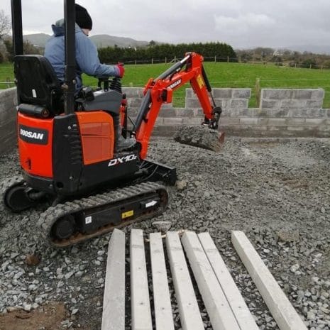 Doosan DX10z Mini Digger-2 A person operates a Doosan DX10z Mini Digger on gravel near a brick wall and concrete beams—ideal for those seeking tool hire Newry options for compact construction work.