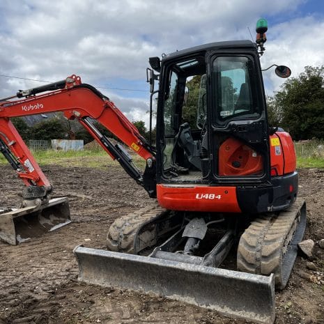 Kubota U48-4-2 A Kubota U48-4 mini excavator, available for tool hire in Newry, is parked on a dirt construction site with its arm and bucket on the ground. Trees and a van are visible in the background.