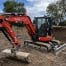 A Kubota U48-4 mini excavator, available for tool hire Newry, is parked on a dirt construction site surrounded by earth mounds and trees under a partly cloudy sky.