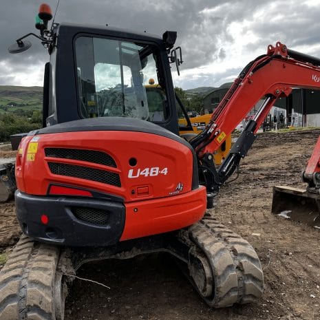 Kubota U48-4-4 A Kubota U48-4 compact excavator with digging bucket stands on a dirt construction site, hills and clouds behind—perfect for tough tasks and available for hire in Newry.