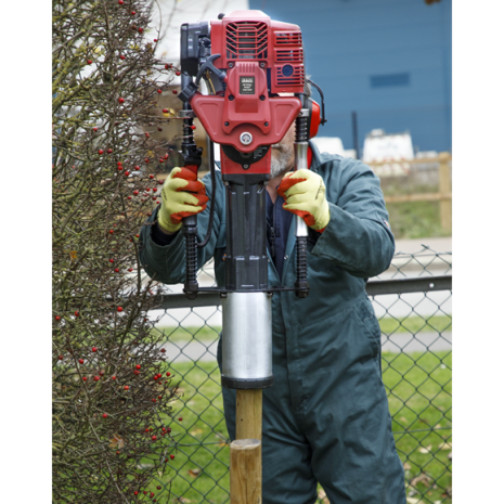 Sealey 2-Stroke Petrol Post Driver 100mm-2 Wearing gloves and overalls, a person uses the Sealey 2-Stroke Petrol Post Driver 100mm from tool hire Newry to drive a wooden stake into the ground beside a fence and bush.