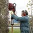 Wearing safety gear, a person uses a Sealey 2-Stroke Petrol Post Driver 100mm to install a wooden stake in the ground near a hedge and chain-link fence.