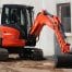 A Kubota U48-4, a compact orange excavator with a black bucket from Tool Hire Newry, is parked on a building site next to a beige building with windows.