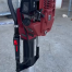The Sealey 2-Stroke Petrol Post Driver 100mm, featuring a red body with a black handle and control buttons, stands upright on concrete and is available for tool hire in Newry.