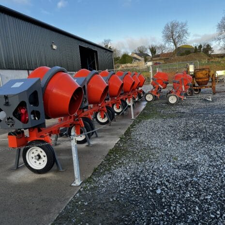 Cargo Tow Behind Mixer-2 A row of Cargo Tow Behind Mixers for tool hire Newry is lined up outside a metal industrial building on a concrete and gravel yard, with trees and houses visible in the background.