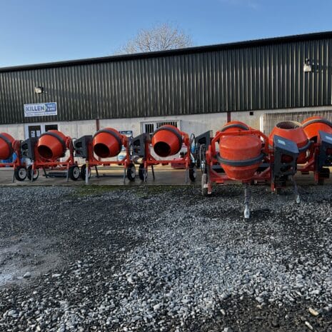 Cargo Tow Behind Mixer-3 A row of red Cargo Tow Behind Mixers, available for tool hire Newry, is lined up outside a grey industrial building on a gravel car park.