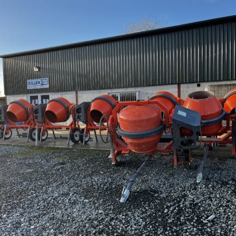 Cargo Tow Behind Mixer-4 A row of red Cargo Tow Behind Mixers, available for tool hire Newry, is lined up outside a metal industrial building labelled "Killen" on a clear day.