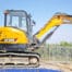 A yellow SANY SY50U compact excavator is parked on a construction site near a white metal fence with greenery in the background—perfect for tool hire Newry projects.