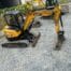 A SANY SY50U mini excavator in yellow, available for tool hire Newry, is parked on gravel near a stone wall, with a Komatsu vehicle seen in the background.