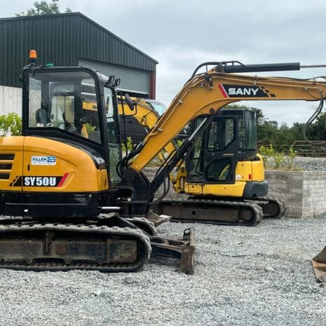 SANY SY50U-5 Two SANY SY50U compact excavators, available for tool hire Newry, are parked on gravel near a farm building with trees and stone walls in the background.