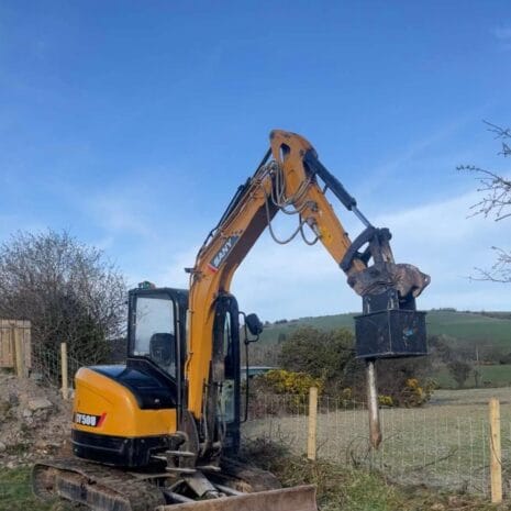 SANY SY50U-2 A SANY SY50U mini digger from Tool Hire Newry lifts a bucket of soil near a wooden fence, with open fields and hills in the background beneath a clear blue sky.