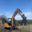 A SANY SY50U mini digger from Tool Hire Newry lifts a bucket of soil near a wooden fence, with open fields and hills in the background beneath a clear blue sky.